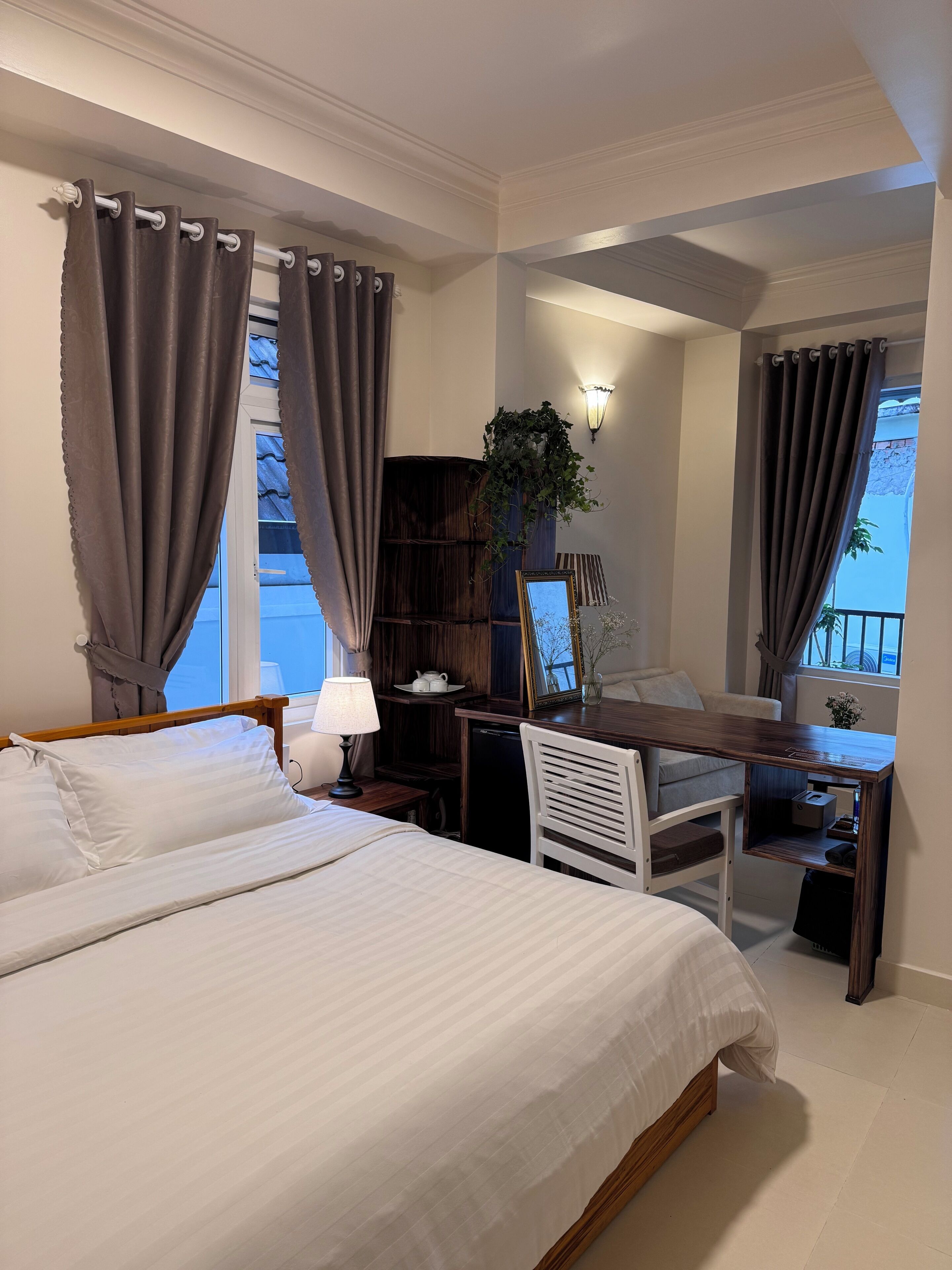 A bedroom with a bed, desk, chair, lamp, and a view of the outside.