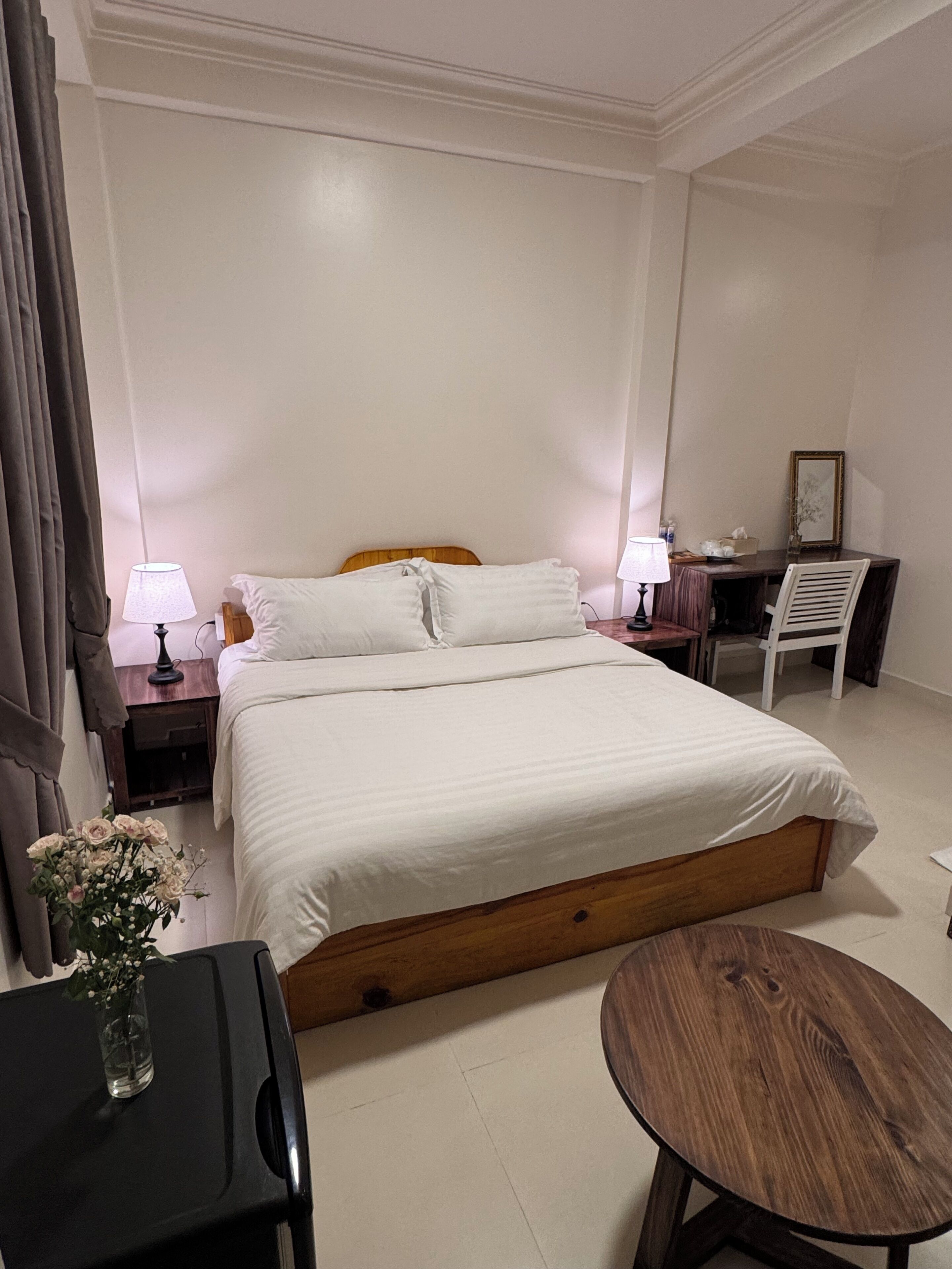 Un dormitorio con cama, mesitas de noche, un escritorio, una silla y una pequeña mesa con un jarrón de flores.