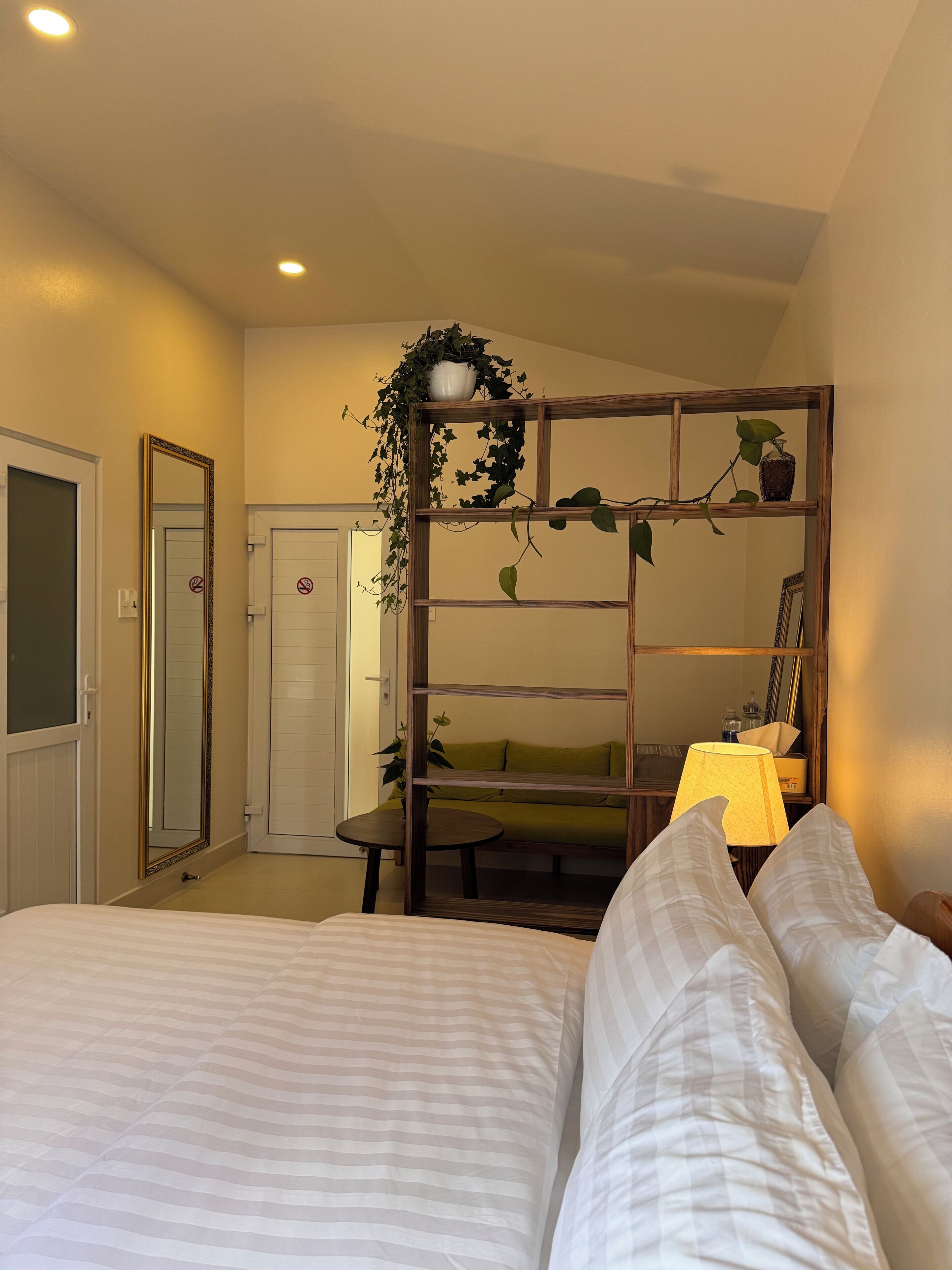 A hotel room with a bed, a small table, a chair, a mirror, and a shelf with plants.