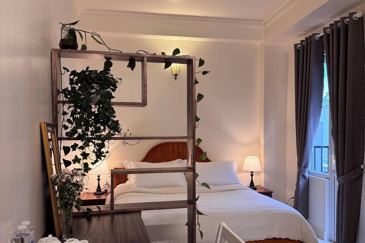 A hotel room with a wooden desk, a bed with white linens, a bedside table with a lamp, and a window with curtains.