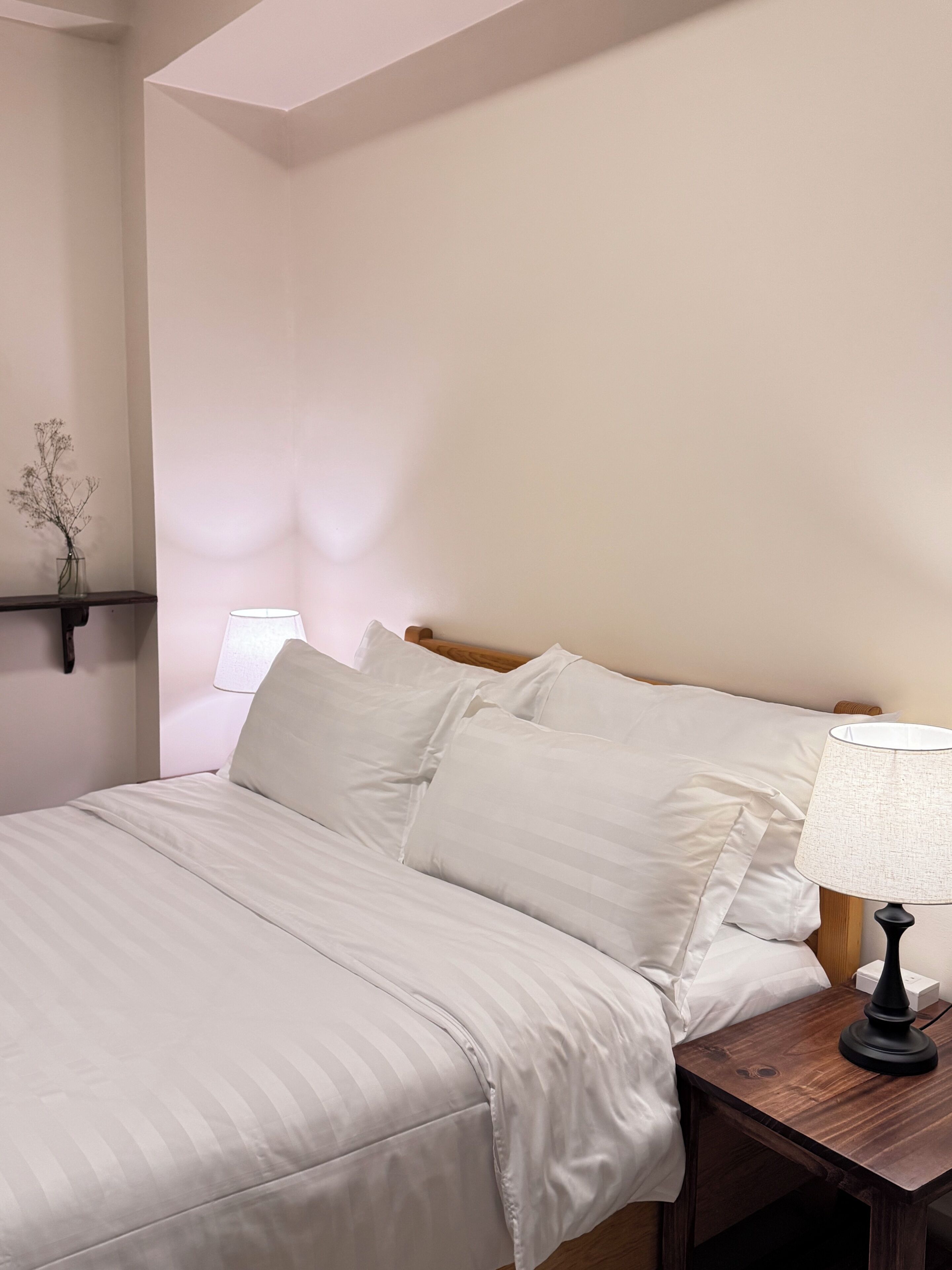A neatly made bed with white bedding, a bedside table with a lamp, and a small vase with dried flowers on a shelf.