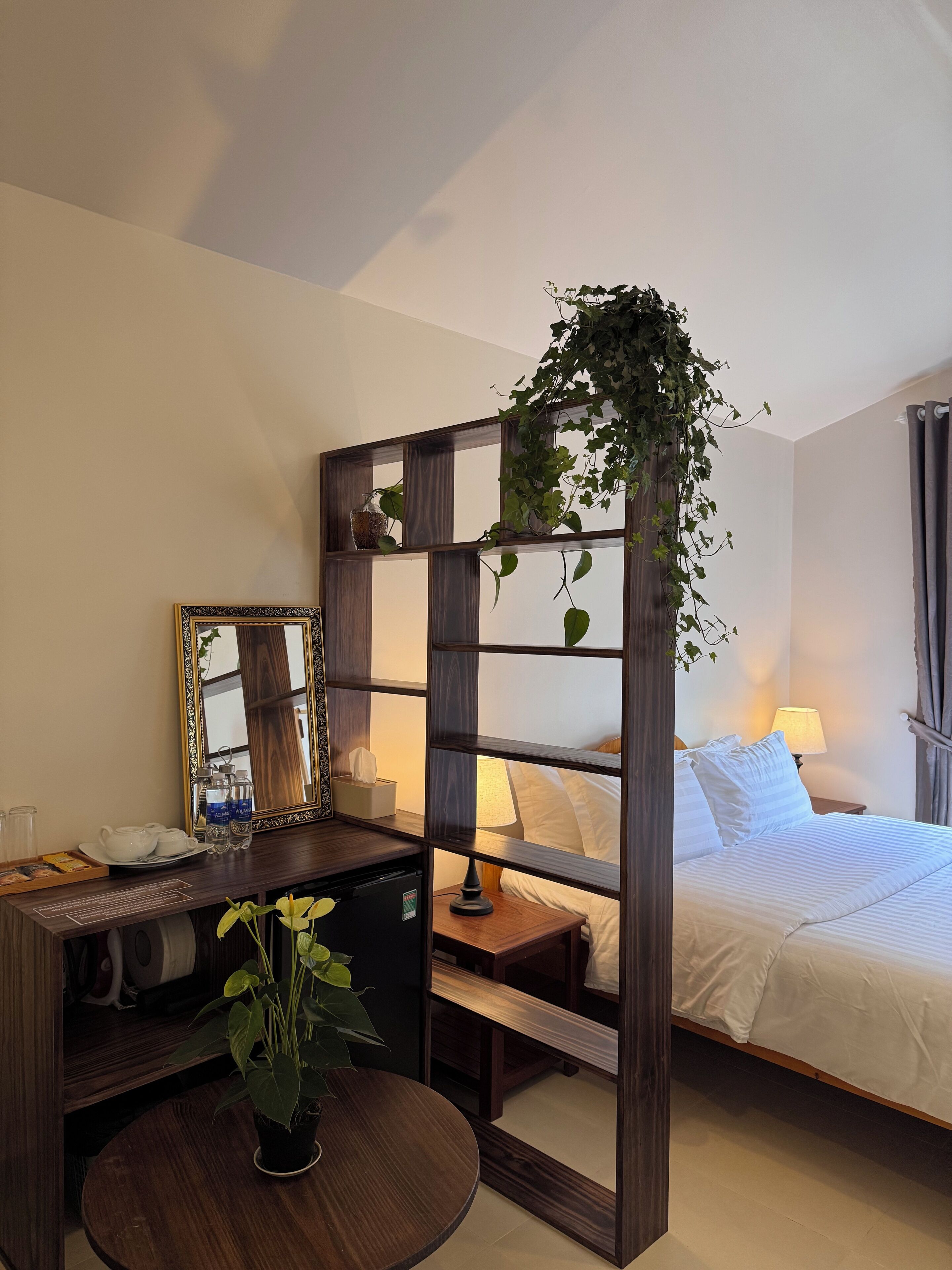 Una habitación de hotel con cama, mesita de noche, lámpara y escritorio con espejo, tetera y botellas de agua.