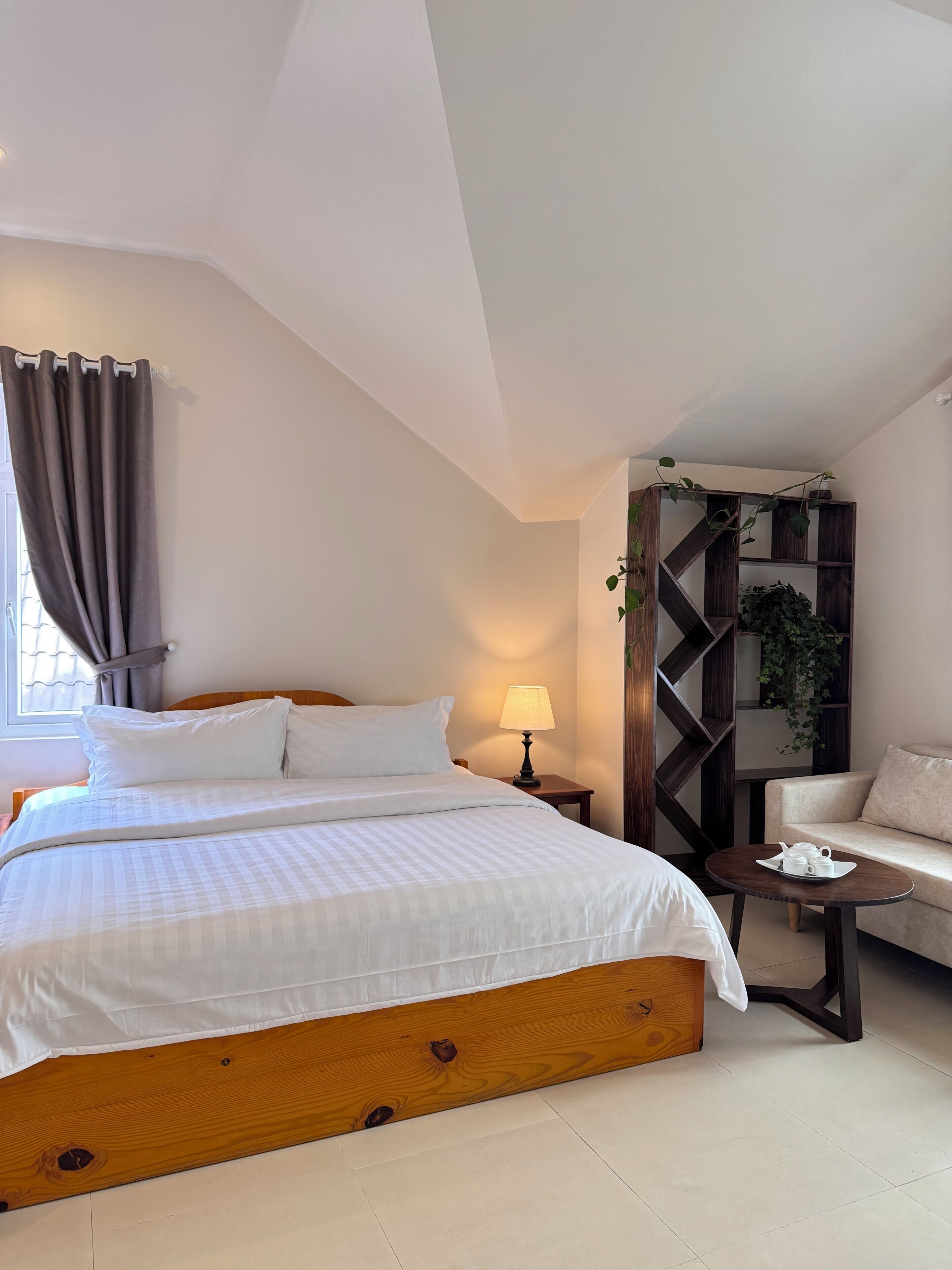 A bedroom with a large bed, a bedside table with a lamp, a wooden headboard, a sofa, and a plant shelf.