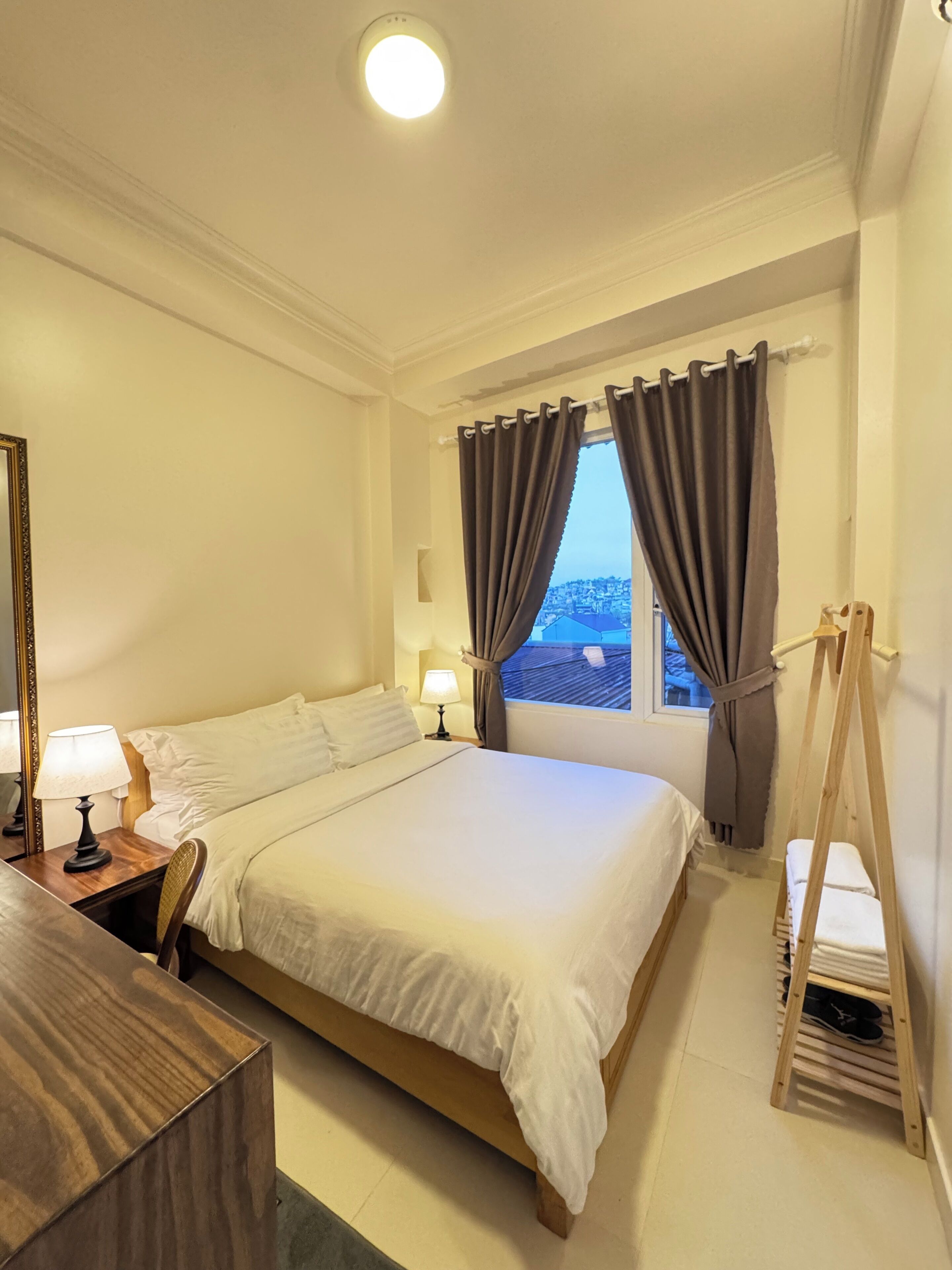 A hotel room with a bed, bedside tables, a mirror, a window with curtains, and a wooden ladder.