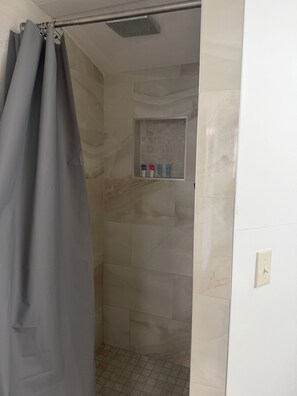 A shower area with a tiled wall, a showerhead, a curtain, and a shelf with toiletries.