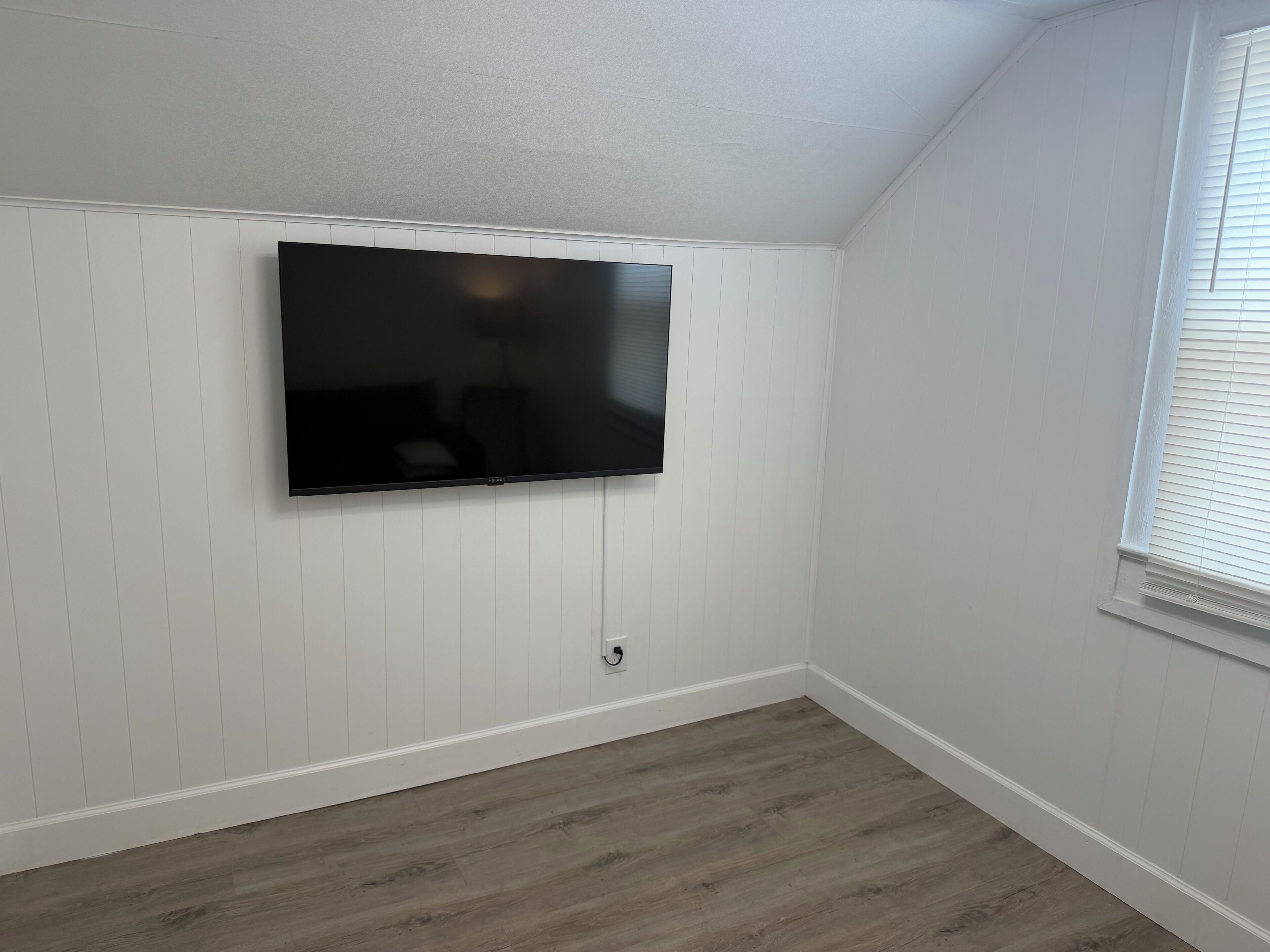Une pièce dotée d’un téléviseur à écran plat fixé au mur, de murs recouverts de panneaux blancs et d’un plancher en bois.
