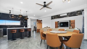 A modern kitchen with a central island, bar stools, a dining area with a round table and chairs, a ceiling fan, and a stone accent wall. - Ultra Modern Luxury Retreat with Pool in Northcrest Estate, sleeps 9 (Berrimah)