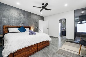 A modern bedroom with a wooden bed, a ceiling fan, and a view into the kitchen.