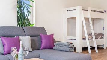Une pièce avec un canapé gris, un lit superposé blanc, une table en bois ornée d’un vase et une grande plante.
