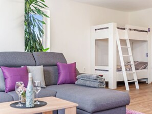 A room with a grey sofa, a white bunk bed, a wooden table with a vase, and a large plant.