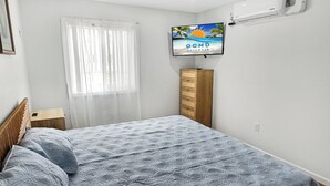 Ein Schlafzimmer mit einem Bett, einem Fernseher an der Wand, einem Fenster mit durchsichtigen Vorhängen und einer Holzkommode.