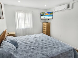 Ein Schlafzimmer mit einem Bett, einem Fernseher an der Wand, einem Fenster mit durchsichtigen Vorhängen und einer Holzkommode.