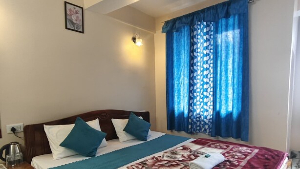 A hotel room with a bed, a nightstand, a green mat, a floral bedspread, and blue curtains. - REJOICE HOMESTAY  (Darjeeling)