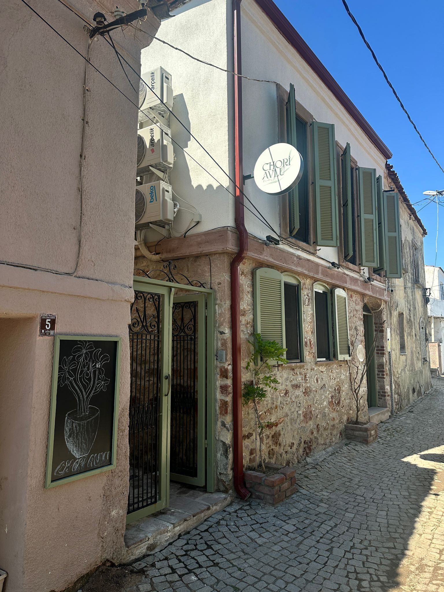 Un edificio con un letrero que dice 'Chori Avlu', fachada de piedra, calle adoquinada y una reja metálica.