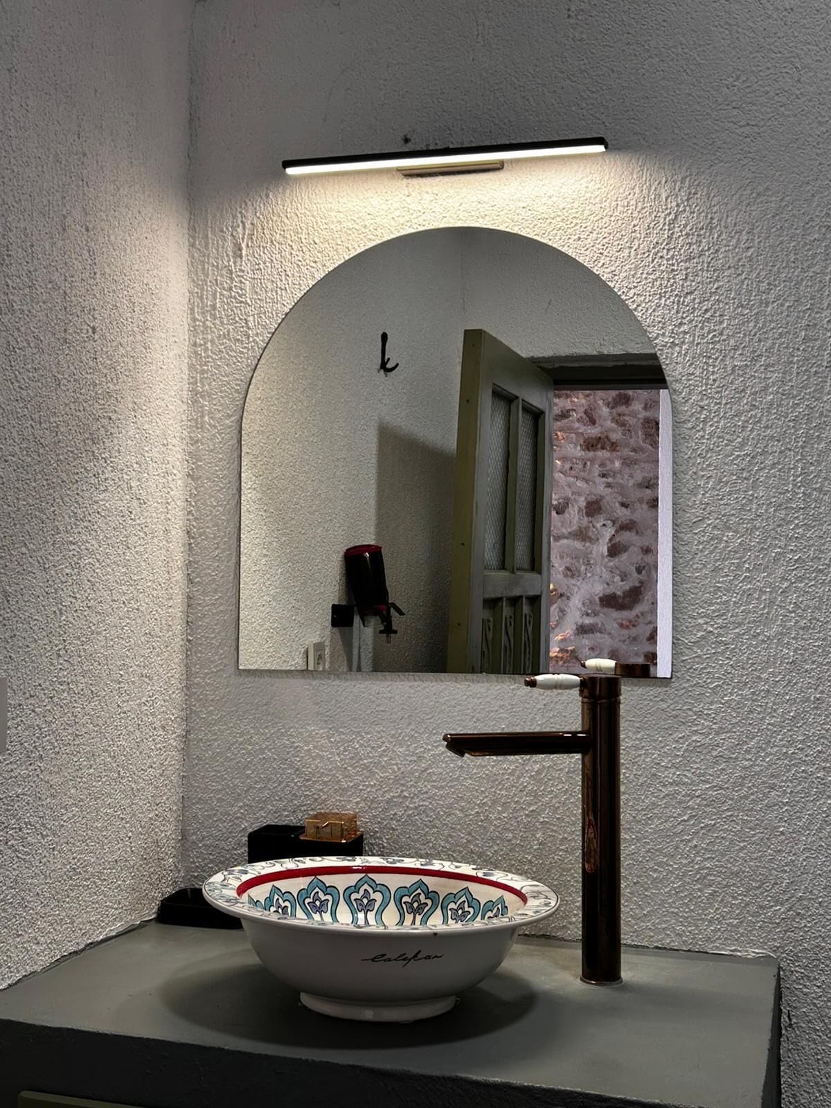 Een badkamer met een witte, geribbelde muur, een spiegel met een gebogen omlijsting, een witte wastafel met een kom met rood en blauw patroon, en een zwarte kraan.
