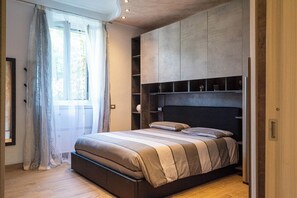 A bedroom with a bed, built-in shelving, a window with curtains, and a wooden floor.