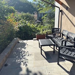 Un patio con mobiliario exterior, suelo de piedra y vistas a una ladera con árboles y edificios.