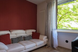 A living room with a white sofa, red cushions, patterned pillows, a radiator, and a window with curtains.