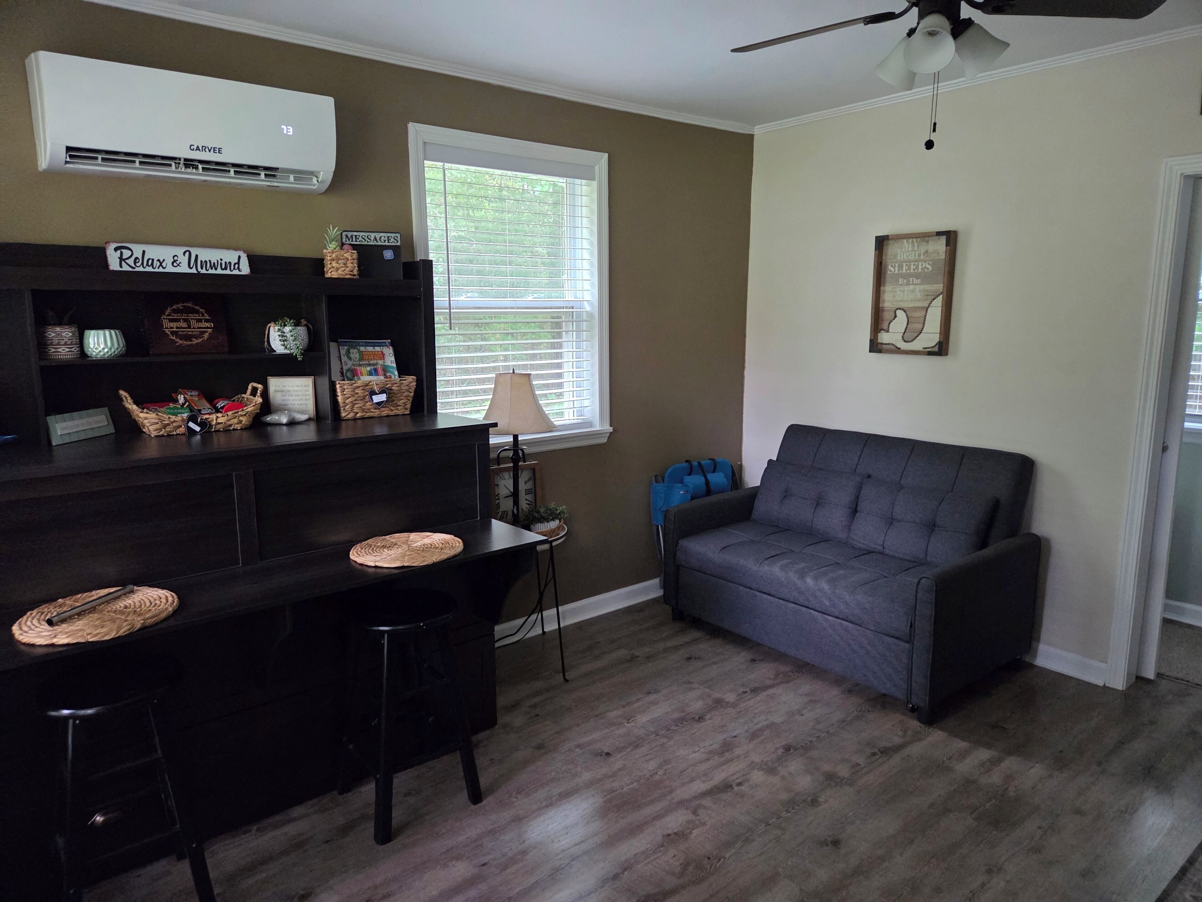 A living room with a dark-colored sofa, a small table with chairs, a wall-mounted air conditioner, and a window with blinds.