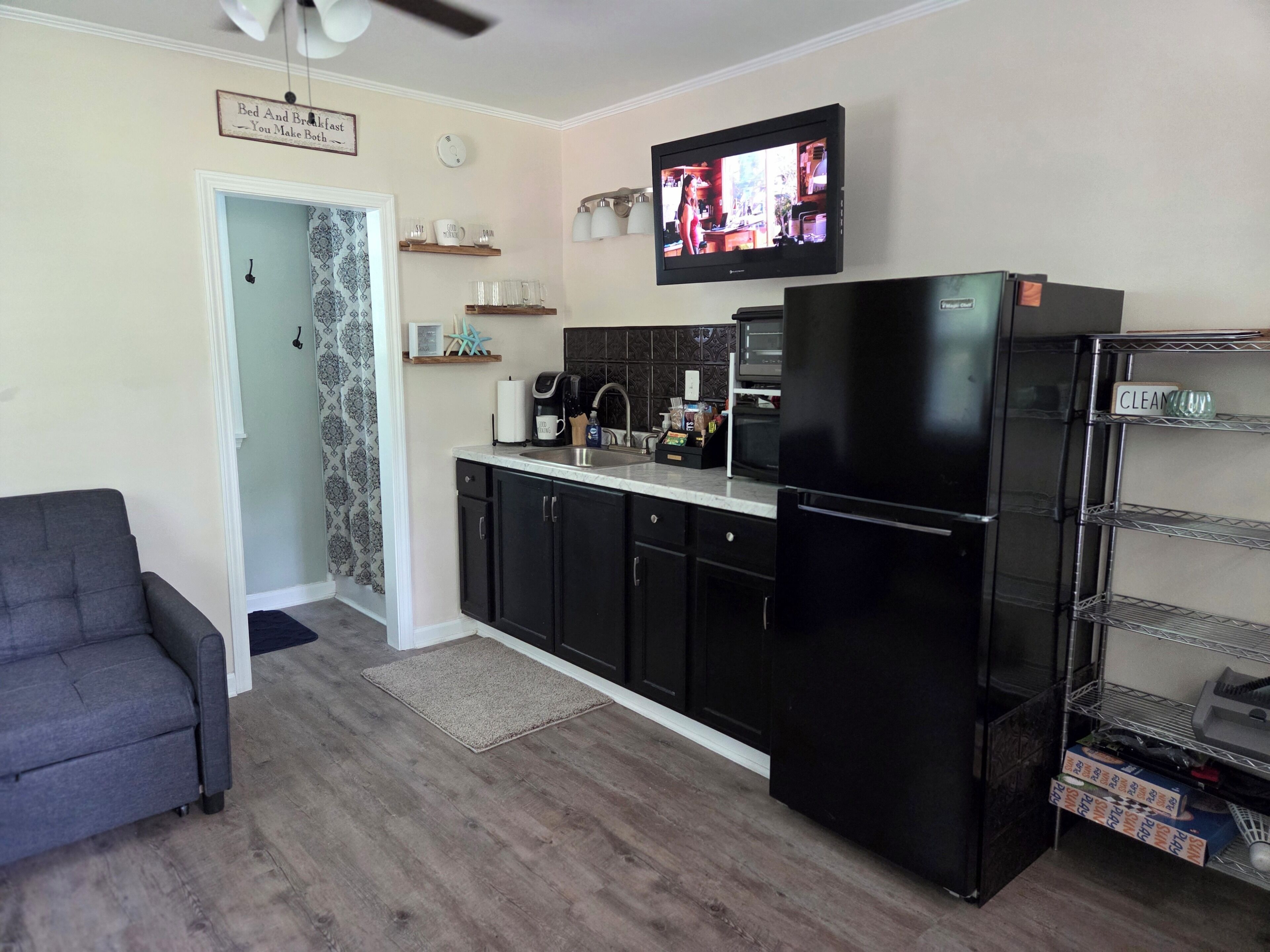 A compact kitchen with a refrigerator, microwave, and coffee maker. There's a TV mounted above the counter and a gray sofa in the living area.