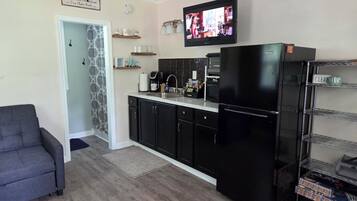 A compact kitchen with a refrigerator, microwave, and coffee maker. There's a TV mounted above the counter and a gray sofa in the living area.