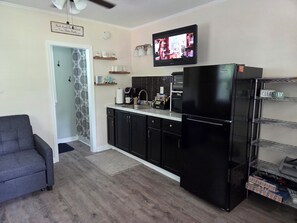 A compact kitchen with a refrigerator, microwave, and coffee maker. There's a TV mounted above the counter and a gray sofa in the living area. - Magnolia Meadows (Hampstead)