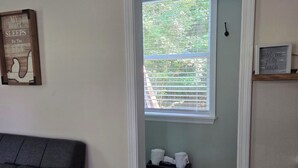 A room with a grey couch, a window with blinds, a small shelf with a glass, and a door leading to another room. - Magnolia Meadows (Hampstead)