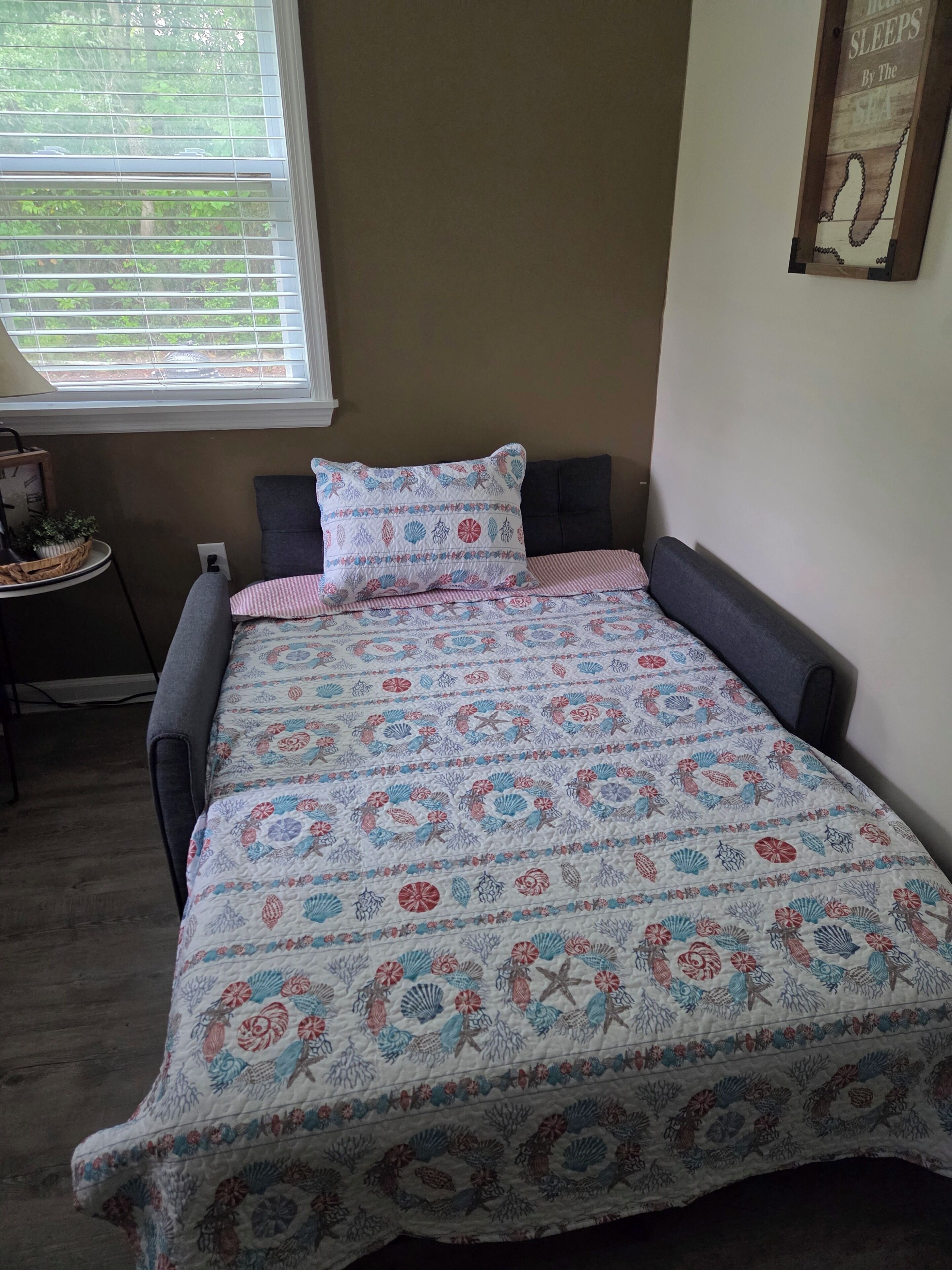 Un dormitorio con cama, ventana con persianas, una mesita con una planta y un letrero en la pared.