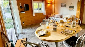 Interior de una cabaña de madera con una mesa de comedor preparada para dos personas, un sofá y un televisor montado en la pared.