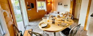 Interior de una cabaña de madera con una mesa de comedor preparada para dos personas, un sofá y un televisor montado en la pared.