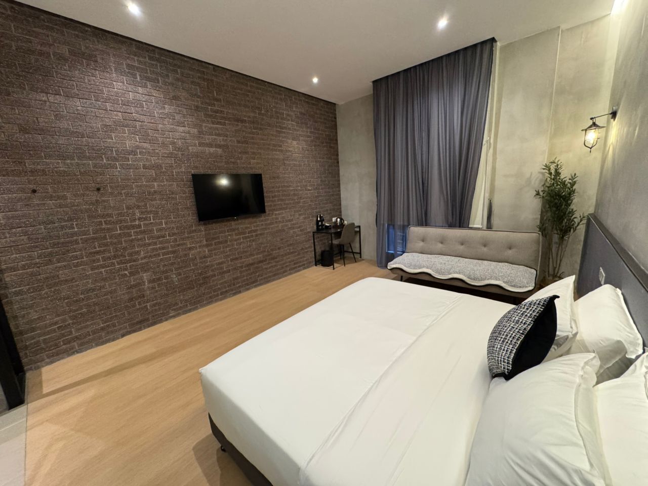 A modern bedroom with a brick wall, a flat-screen TV, a bed with white linens, a desk, and a chair.