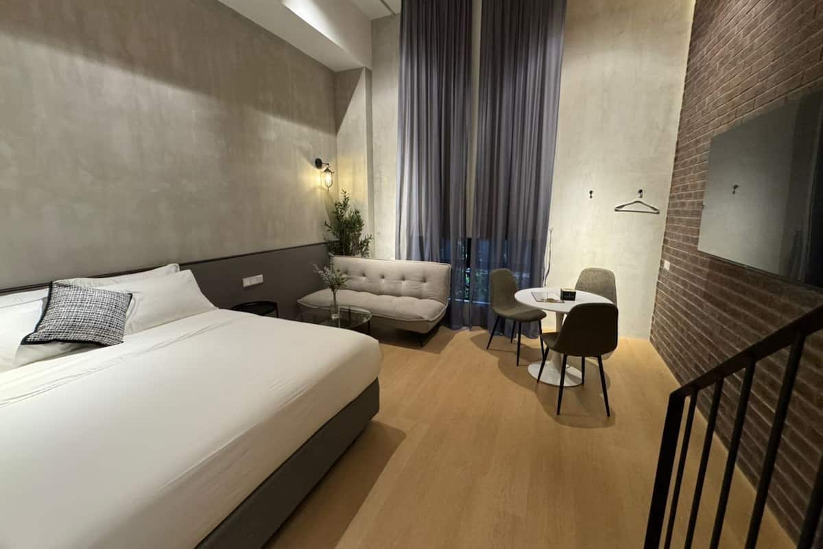 Una habitación de hotel moderna con una cama grande, un sofá, una mesita pequeña y una silla. Hay una ventana con cortinas, una lámpara de pared y una pared decorada con ladrillos.