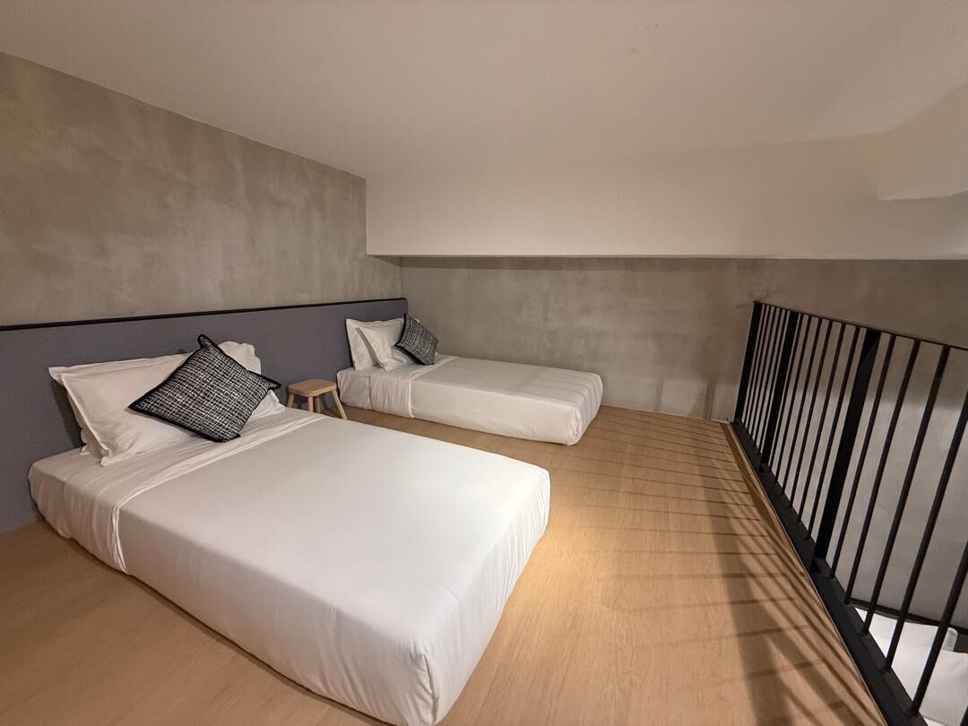 A hotel room with two beds, a small stool, a balcony with a black railing, and a concrete wall.