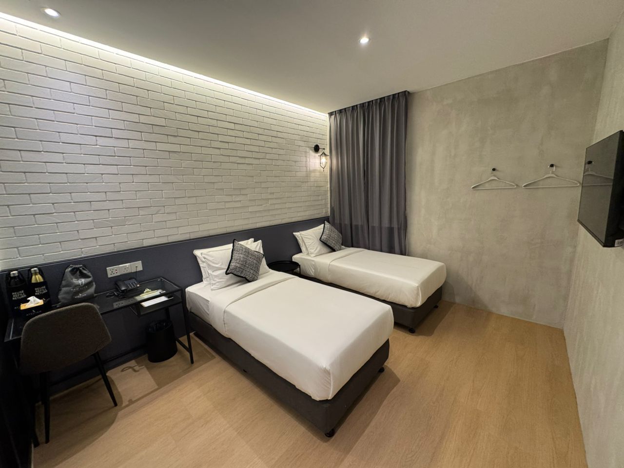 A modern hotel room with two single beds, a desk with a chair, a small trash bin, and a wall-mounted TV.