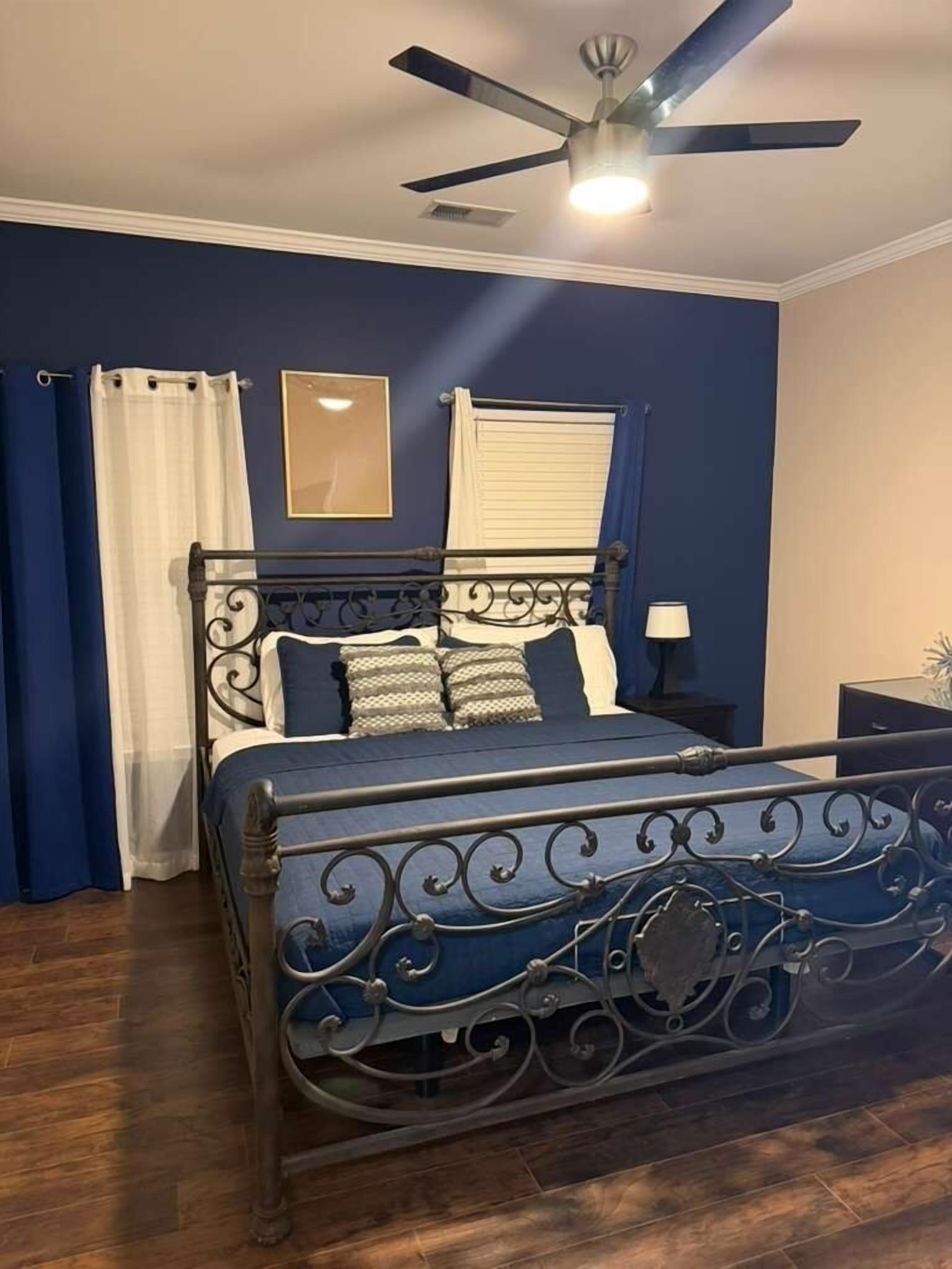 Une chambre à coucher avec un lit, un ventilateur de plafond, une table de chevet et une fenêtre avec des rideaux.
