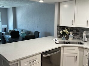 A modern kitchen with white cabinets, a dishwasher, and a countertop. There is a dining area with a sofa and a laptop on the table. - Ocean Magic 2/2 Condo on the Beach Great Ocean Views Pool 4G (Daytona Beach)