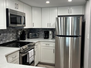 A modern kitchen with white cabinets, stainless steel appliances, a microwave, toaster oven, coffee maker, and a refrigerator. - Ocean Magic 2/2 Condo on the Beach Great Ocean Views Pool 4G (Daytona Beach)
