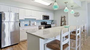 A modern kitchen with white cabinets, stainless steel appliances, a marble countertop, and wicker chairs.