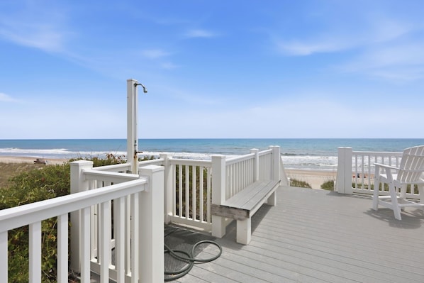 A wooden deck with white railings, a bench, and a chair overlooking a beach.