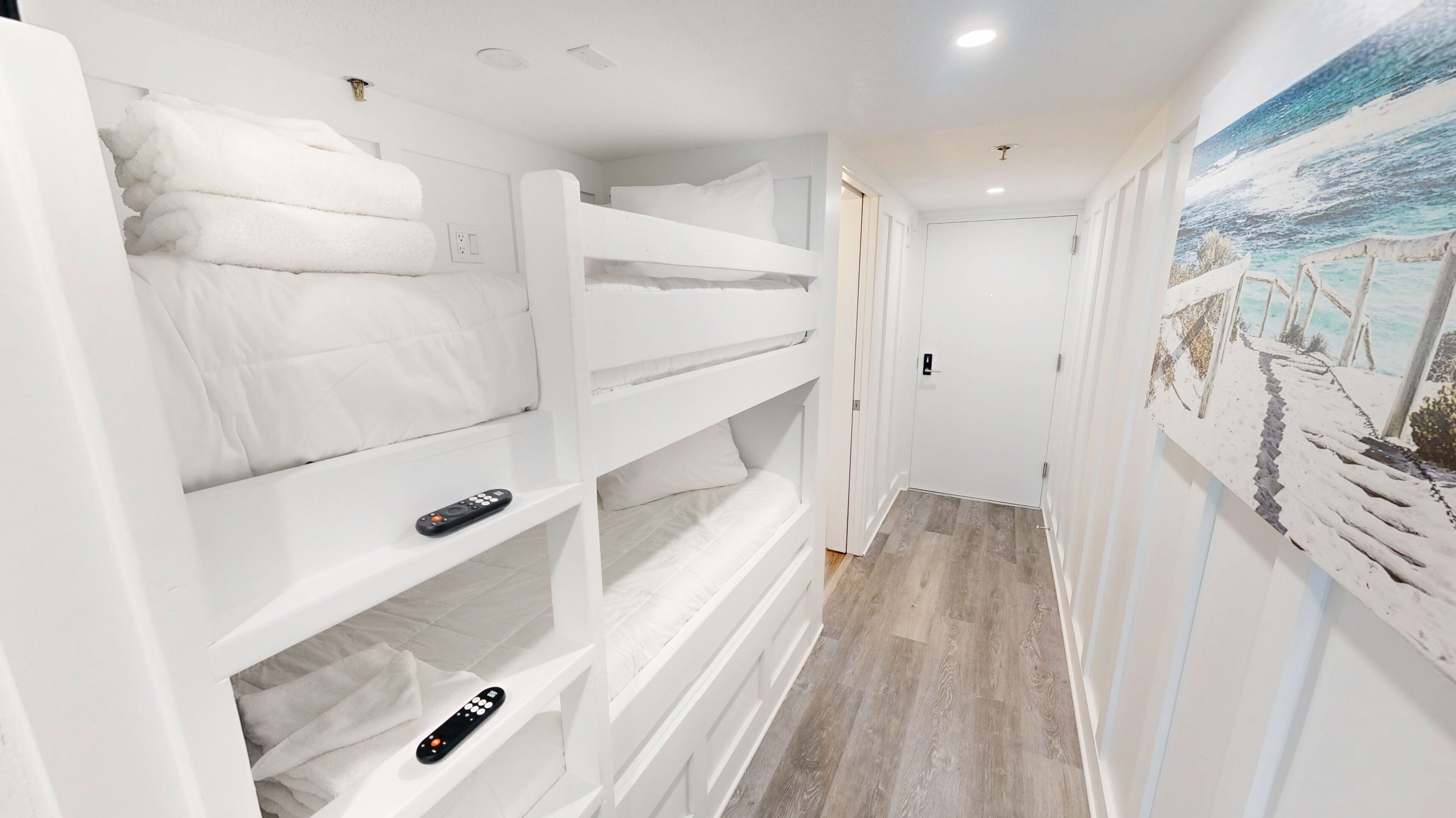 Habitación con literas, un mando a distancia en la litera inferior, una puerta y un cuadro de una escena costera en la pared.