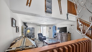 A high-ceilinged living room with a stone fireplace, a large armchair, and a wooden staircase. - Charming Home Near Lake Winnipesaukee Beach Access 3 Bedroom Home (Moultonborough)