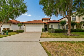 A residential house with a red-tiled roof, a garage, and a front yard with trees and shrubs. - Serene Villa with Heated Pool & Arcade Near Disney (Clermont)