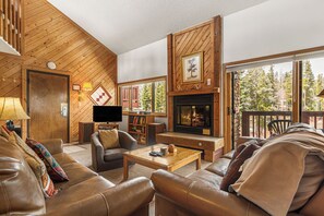 Ein gemütliches Wohnzimmer mit Ledersofas, einem Kamin, einem Fernseher und einem großen Fenster mit Blick auf Bäume.