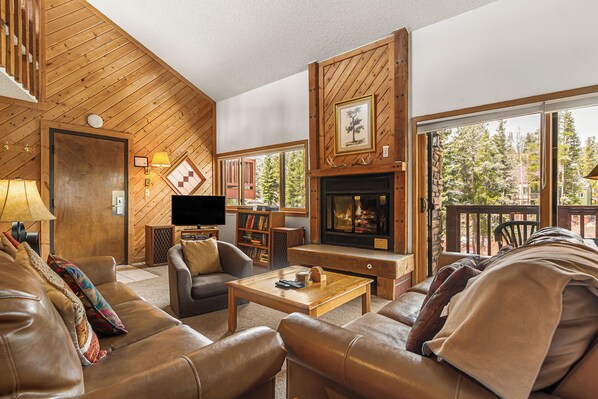 A cozy living room with leather sofas, a fireplace, a television, and a large window with a view of trees. - Ski-In Condo | Loft + Fireplace | Near Main St (Breckenridge)