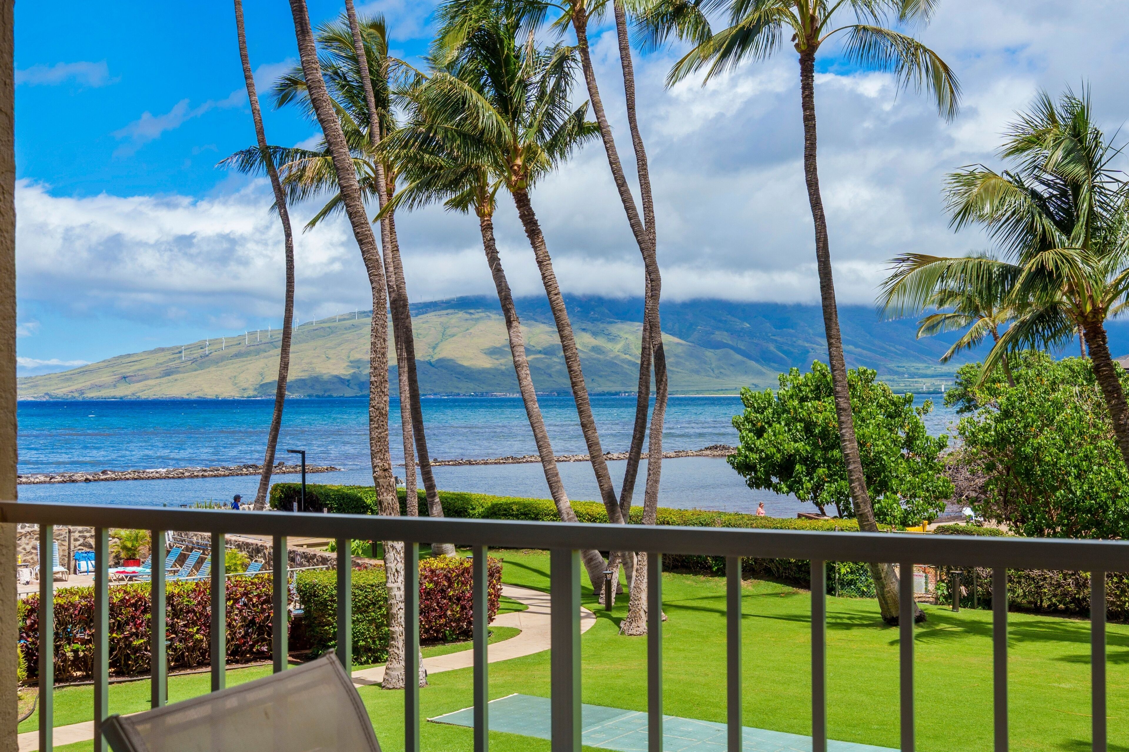 A view from a balcony overlooking a beach with palm trees, a clear sky, and distant mountains.