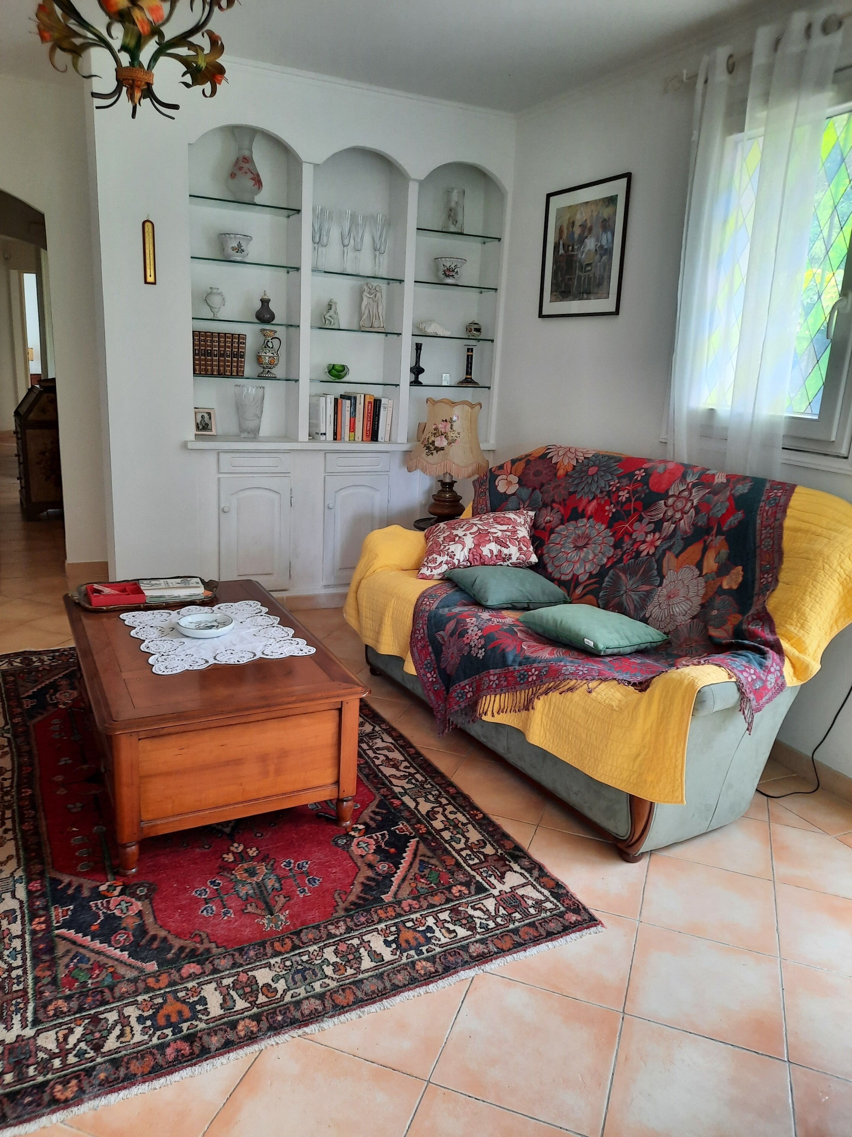 Een woonkamer met een bank, een salontafel, een kamerkleed met patroon en een boekenkast met decoratieve objecten.