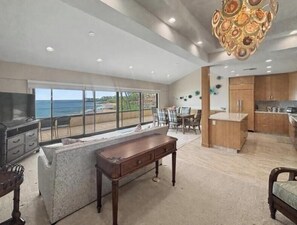 Un amplio salón con un gran ventanal con vistas al océano, un comedor con mesa y sillas, y una cocina con armarios de madera.