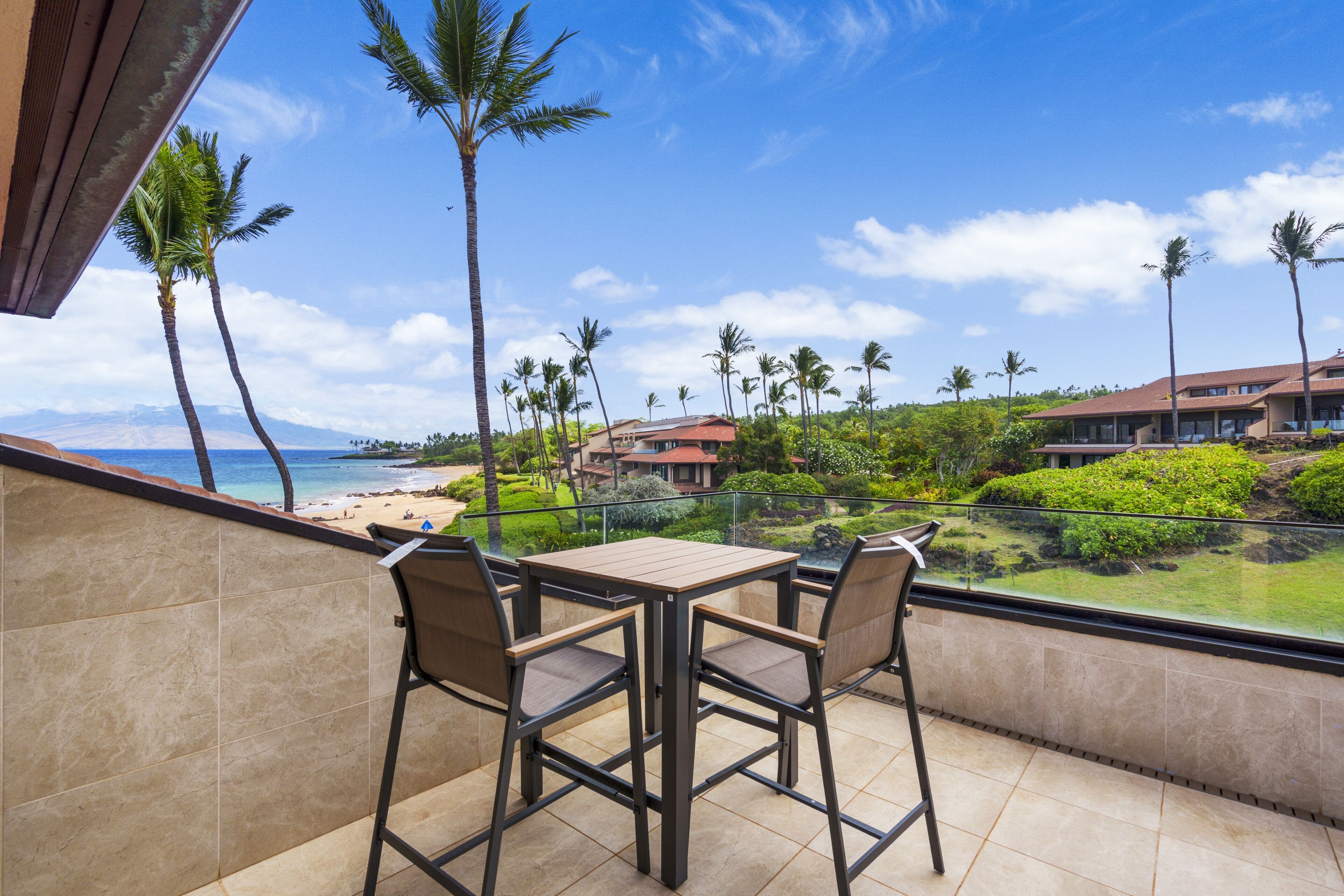 A balcony with a table and chairs overlooking a beach and resort buildings.