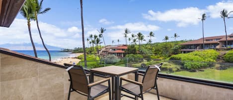 A balcony with a table and chairs overlooking a beach and resort buildings.