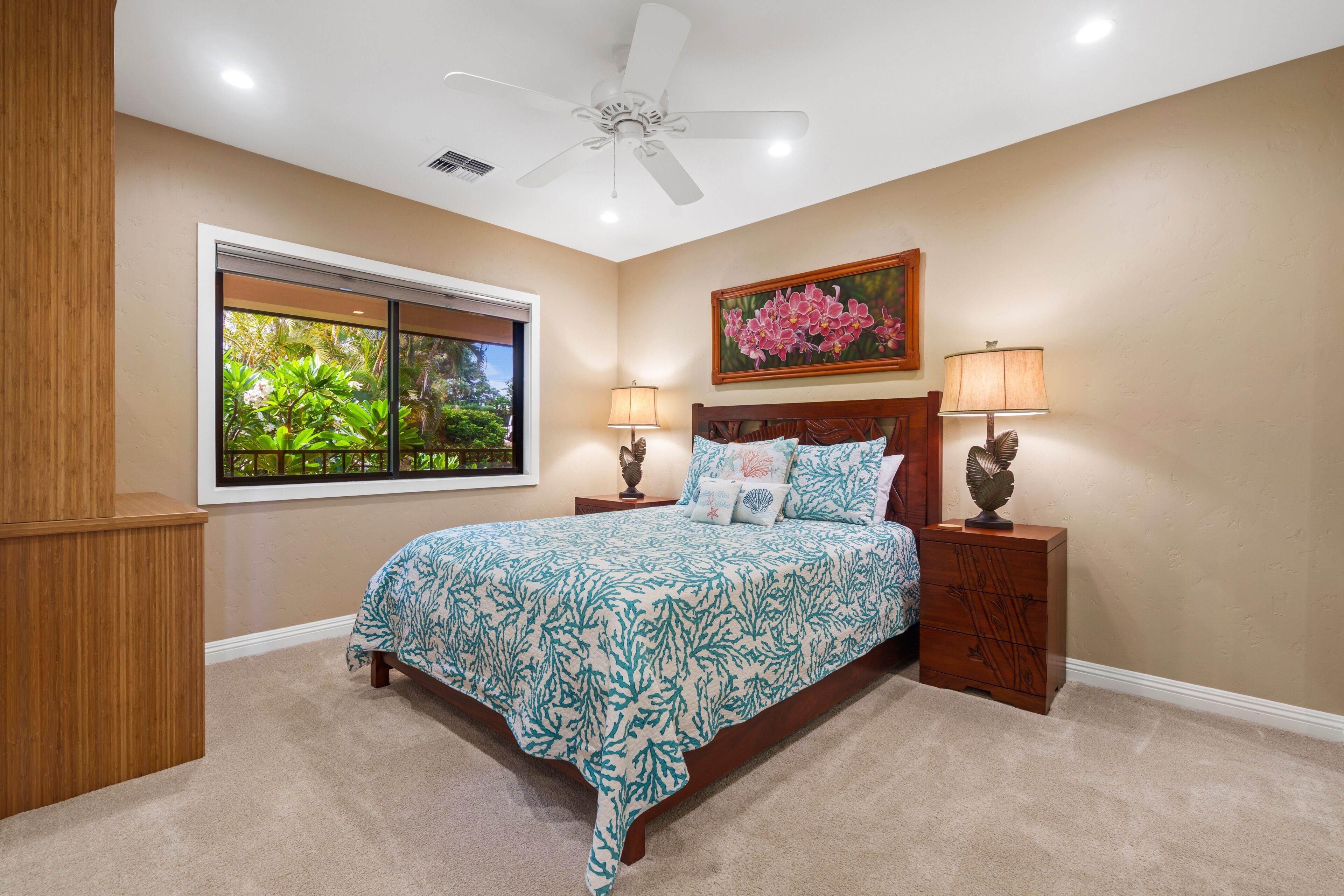 Quarto com cama, duas mesas de cabeceira, ventilador de teto, janela com vista para o verde e um quadro na parede.